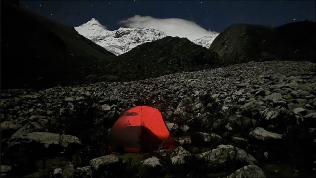 Ein rot beleuchtetes Zelt umgeben von großen Steinblöcken vor einer Berg-Silhouette teilweise mit leuchtendem Schnee unter einem Sternenhimmel in Peru