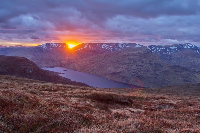 Spektakulärer Sonnenuntergang in Schottland, bei dem die rote Sonne gerade noch zwischen den Bergen hervorschaut.
