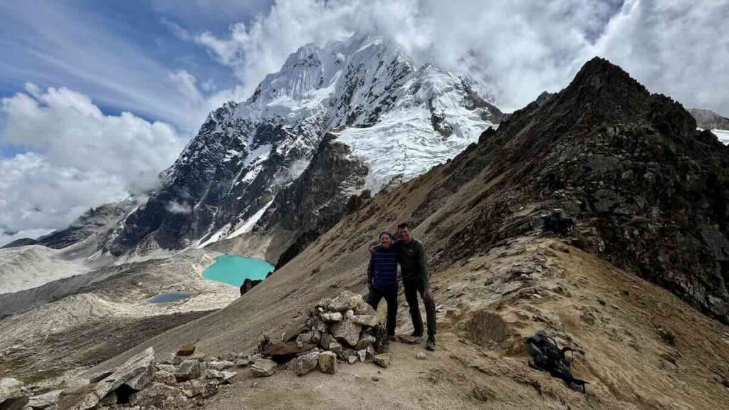Zwei Wanderer auf einem Pass über einem Türkisen Bergsee mit einem schneebedecktem Gipfel im Hintergrund in Peru